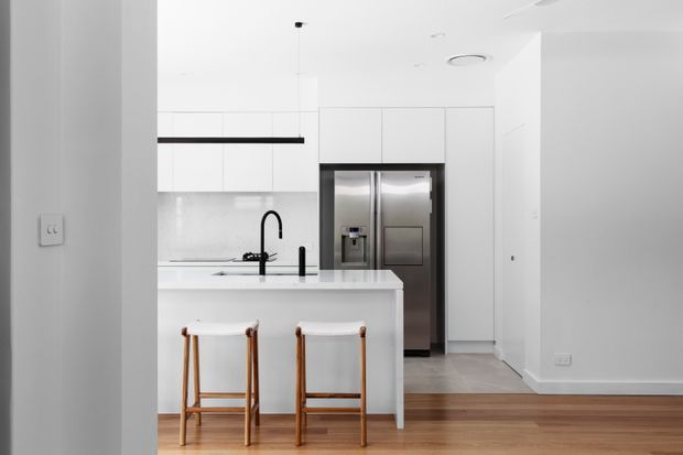 Bright contemporary white kitchen viewed from an adjoining room, featuring a white island with two timber-framed stools, integrated white cabinetry, a pale stone benchtop and splashback, black linear pendant lighting and tapware, stainless-steel French-door refrigeration, and warm timber flooring.