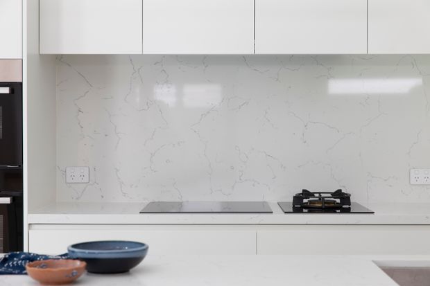 Bright contemporary kitchen with white flat-panel cabinetry, a white veined stone splashback and benchtop, integrated induction cooktop, and a black gas cooktop. A white island or counter occupies the foreground with simple ceramic bowls, creating a clean, minimal composition.