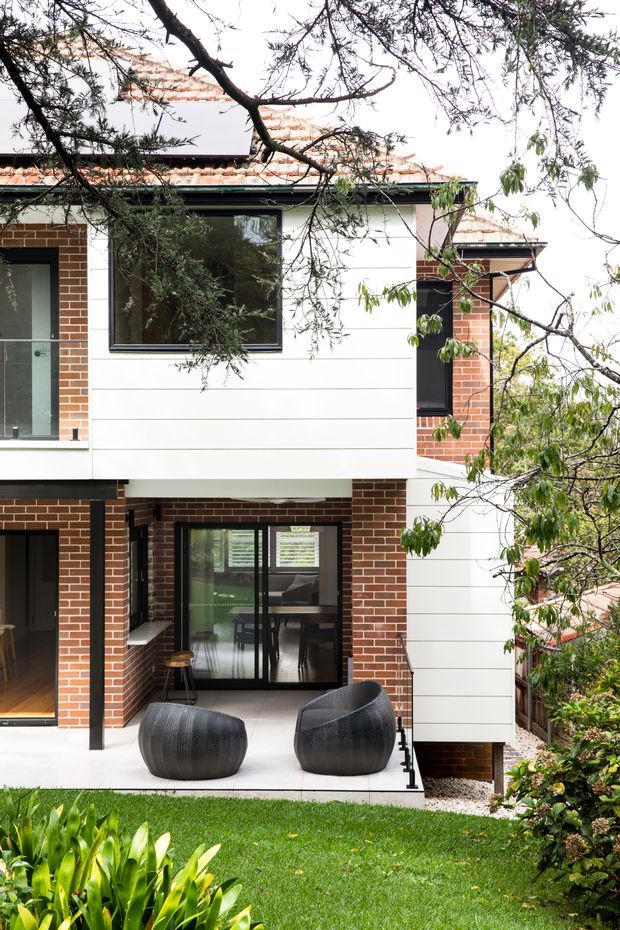 Exterior view of a contemporary two-storey brick and white-clad residence, partially screened by mature trees. The facade combines red brickwork, white horizontal cladding, dark window and door frames, and a tiled pitched roof. A covered ground-floor patio with two dark outdoor lounge chairs opens toward a landscaped lawn and garden.