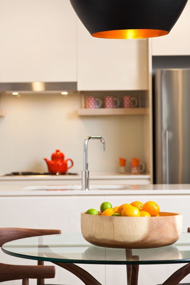 A warmly lit contemporary kitchen viewed from a dining area, featuring white cabinetry, a stainless-steel refrigerator, a pale benchtop with a tall chrome tap, open shelving with patterned mugs, and a large black pendant light with a glowing orange interior. In the foreground, a glass dining table with dark timber chairs holds a wooden bowl of citrus fruit.