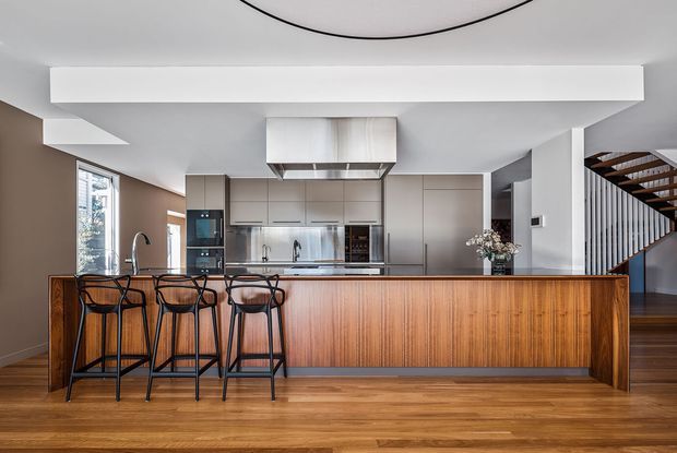 A contemporary open-plan kitchen with a long timber island and integrated breakfast bar, four black metal bar stools, pale cabinetry, stainless-steel appliances, and a large sculptural rangehood. Warm timber flooring and a staircase with dark timber treads are visible at right, while a large circular ceiling light adds a distinctive feature overhead.