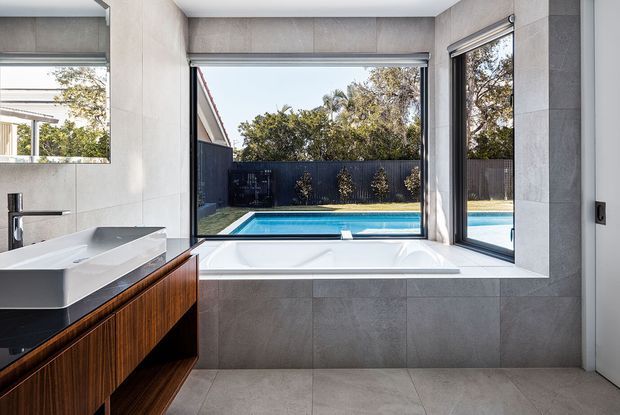 Contemporary bathroom with a freestanding bathtub set into a tiled window alcove overlooking a swimming pool and landscaped courtyard. The room features large-format grey wall and floor tiles, a floating dark timber vanity with a white integrated basin, brushed metal tapware, and expansive black-framed windows.