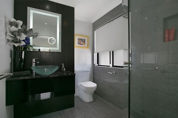 Contemporary bathroom featuring a glossy dark built-in vanity with a transparent vessel basin, large framed mirror, wall-mounted tapware, toilet, and a glass-enclosed shower. Grey stone-look wall and floor finishes create a sleek monochromatic scheme, with a window fitted with a roller blind and subtle floral styling.