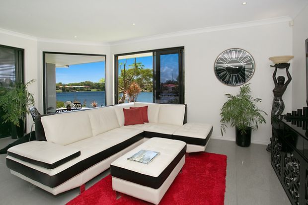 Bright contemporary living room with a large white and black sectional sofa, matching ottoman, vivid red area rug, potted plants, decorative wall clock, and dark entertainment cabinetry. Large windows and glazed doors overlook a calm waterfront setting.