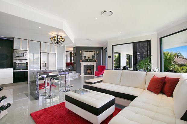 Bright open-plan living and kitchen area with a large white sectional sofa, black-and-white ottoman, red rug and cushions, a marble-look kitchen island with bar stools, and extensive glazing opening toward an outdoor view. The room includes a decorative fireplace wall and chandelier-style lighting, with a polished contemporary fit-out.