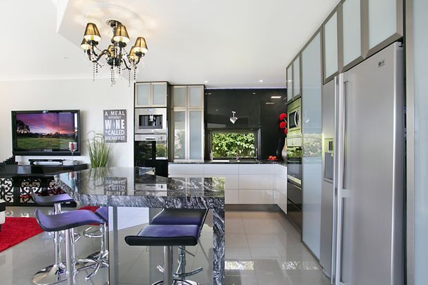 Bright open-plan kitchen and dining area with a polished dark stone island and breakfast bar, integrated stainless-steel appliances, glossy white cabinetry, and a black glass splashback. A formal dining table, decorative chandelier, television, and garden view through the rear window are also visible.