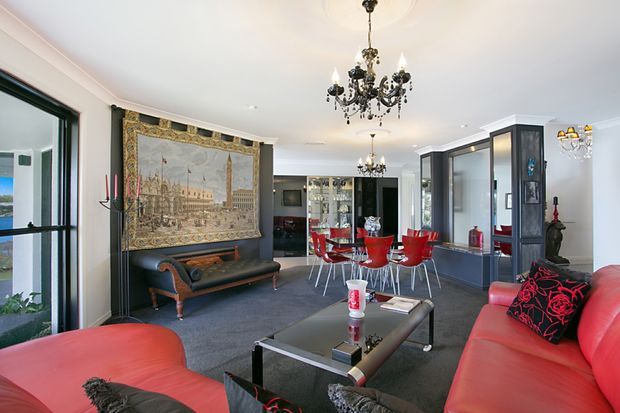 A spacious, elegant living and dining room with a dramatic black, white, and red palette. The room features red leather sofas, a black coffee table, a red dining table with matching chairs, dark built-in cabinetry and display units, a large framed tapestry or artwork, and ornate crystal chandeliers. Large glazed openings provide views to the exterior, while decorative furnishings and traditional detailing create a refined, eclectic atmosphere.