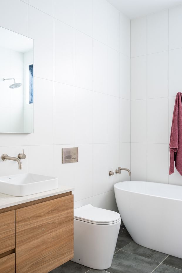 Bright, compact contemporary bathroom with full-height white wall tiling, a wall-mounted mirror, floating timber vanity with white vessel basin, wall-mounted brushed metal tapware, concealed cistern toilet, freestanding white bathtub, and dark grey tiled flooring. A burgundy towel provides the main accent colour.