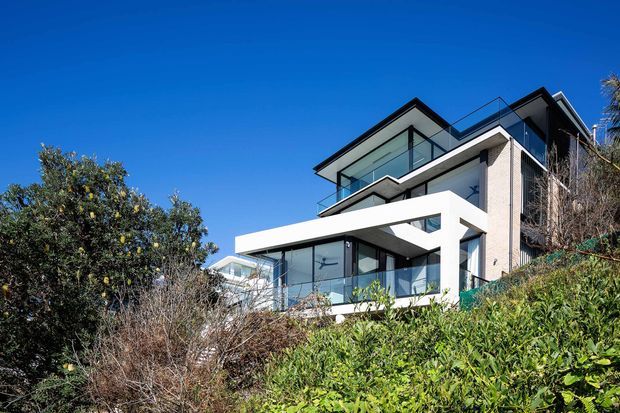 Exterior view of a contemporary multi-storey hillside residence with crisp white rectangular projecting forms, pale masonry walls, extensive floor-to-ceiling glazing, dark window frames, and glass balustrades. Dense landscaped vegetation partially screens the lower level, while the upper levels feature flat roof planes and cantilevered balconies under a clear blue sky.