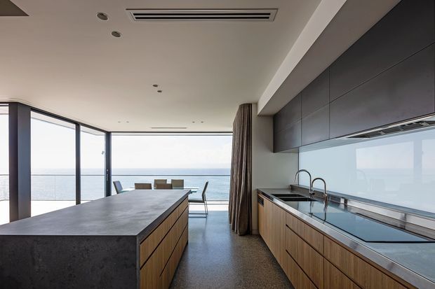 Contemporary open-plan kitchen and dining area with a long dark stone island, timber drawer cabinetry, dark overhead joinery, integrated appliances, and a full-height glazed corner overlooking the sea. A dining table and chairs sit beyond the island beside the expansive ocean-facing glazing.