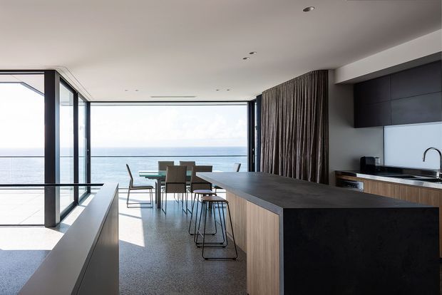 A contemporary open-plan kitchen and dining area with expansive floor-to-ceiling glazing overlooking the ocean. The kitchen features a large dark island with timber cabinetry, a pale splashback, integrated appliances and a minimalist black-framed window wall. A dining table sits beside the glazing, while a textured vertical timber screen separates the kitchen from the adjacent area. The space is bright, refined and strongly connected to the waterfront outlook.
