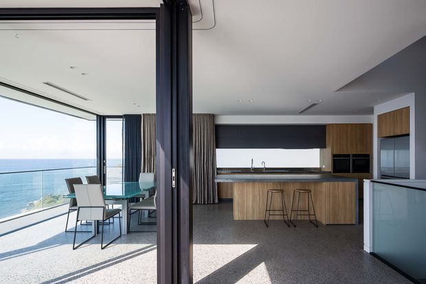 A bright contemporary open-plan kitchen and dining area adjoining a glazed balcony or terrace with expansive ocean views. The kitchen features a wood-grain island and tall cabinetry, dark matte overhead cabinetry, integrated appliances, a pale stone benchtop, and terrazzo-like flooring. A black-framed glazed folding or sliding opening separates the interior from the dining area, while full-height curtains and large windows emphasize the coastal outlook.