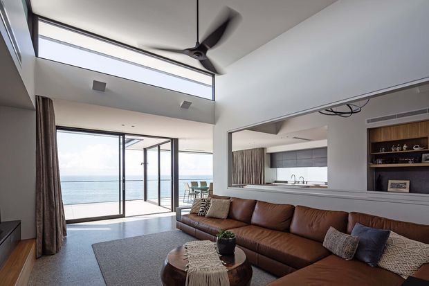 Bright contemporary open-plan living room with a double-height ceiling, ceiling fan, large clerestory glazing, and full-height sliding glass doors opening to a balcony with ocean views. A large brown leather sectional sofa, textured grey rug, timber coffee table, and neutral cushions occupy the foreground. The kitchen is visible through a wide internal opening, with grey cabinetry and a white benchtop. Built-in dark timber shelving and recessed air-conditioning add architectural detail.