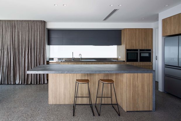 A contemporary kitchen viewed straight on, centred around a large timber-clad island with a dark grey stone benchtop and two black bar stools. The kitchen includes integrated black cabinetry, warm timber overhead and island joinery, a stainless-steel sink and tap, built-in black appliances, and a full-height refrigerator at right. Floor-to-ceiling pleated curtains and a grey carpeted floor contribute to the restrained, neutral palette.