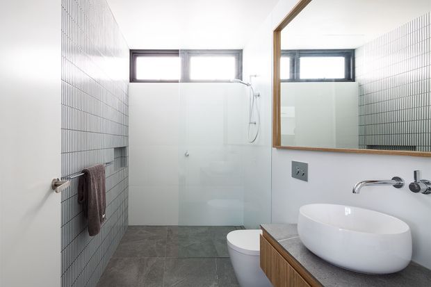 Bright contemporary bathroom with a walk-in shower enclosed by clear glass, freestanding oval vessel basin on timber vanity cabinetry, wall-mounted chrome tapware, toilet, large mirror, small high-set windows, white wall surfaces, vertically gridded grey wall tiles, and dark grey floor tiles.