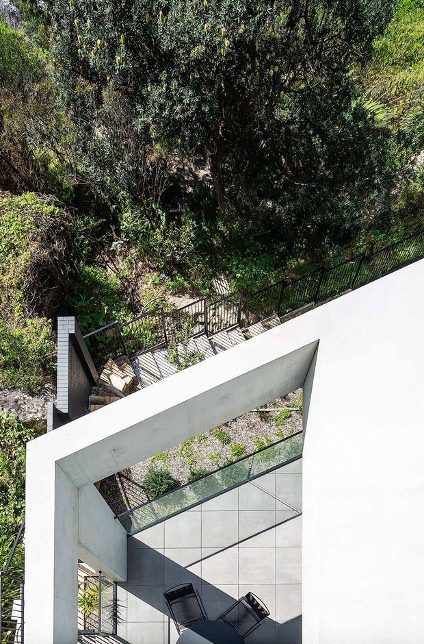 A high-angle exterior architectural view of a contemporary white concrete structure integrated into a steep, densely planted landscape. A sharply framed covered terrace or courtyard occupies the foreground, with large pale tiled surfaces, dark metal railings, outdoor lounge chairs, and a small round table. The building's geometric form contrasts with the surrounding mature greenery and planted roof or garden edge.