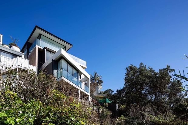 Exterior view of a multi-storey contemporary residence on a sloping, landscaped site. The house features strong angular projecting forms, extensive floor-to-ceiling glazing with glass balustrades, dark vertical cladding, white rendered surfaces, and a low-pitched roof. Dense vegetation partially screens the lower level.