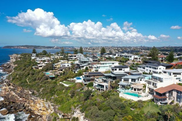 Aerial coastal view of a densely developed residential neighbourhood on a rocky headland, with numerous modern detached homes, terraces, balconies, swimming pools and landscaped gardens overlooking the blue water. A broad bay and urban shoreline are visible in the distance under a bright sky.