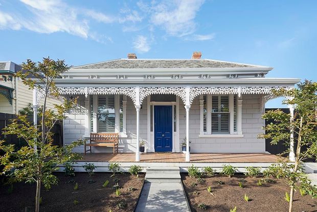 Front elevation of a single-storey heritage-style cottage with pale weatherboard cladding, a low hipped roof, ornate white lacework verandah detailing, decorative brackets and columns, and a vivid blue entry door. A formal front garden and central paved path lead to the raised porch.