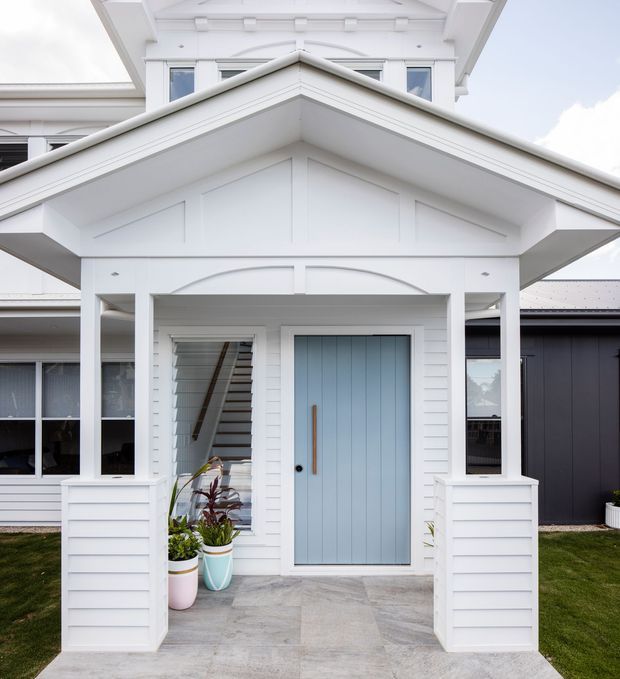 Front exterior view of a bright white weatherboard home with a prominent gabled entry porch, classical trim details, white columns, and a pale blue vertically panelled front door. The entry is approached by a light grey tiled path, with potted plants and glimpses of the stairway and adjoining wings visible through the open porch.