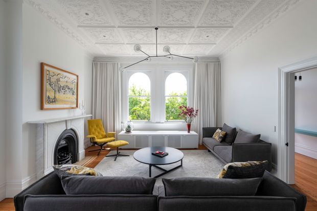 A bright, refined living room with a traditional fireplace, ornate pressed-tin ceiling panels and decorative cornice, tall arched-top windows, and layered contemporary furnishings. Two dark sofas face a round coffee table, while a mustard accent chair sits beside the fireplace. White walls and joinery, pale curtains, timber flooring, and a large light rug create a balanced palette with yellow and floral accents. An adjacent room is visible through the open doorway.