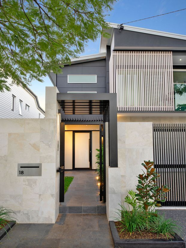 Contemporary multi-storey residential facade viewed from the street, featuring dark grey rendered walls, pale stone boundary walls, vertical privacy screening, a covered entry walkway, and a black-framed glazed front door. The landscaped frontage includes planted garden beds and a paved path, with a neighbouring weatherboard house visible to the side.