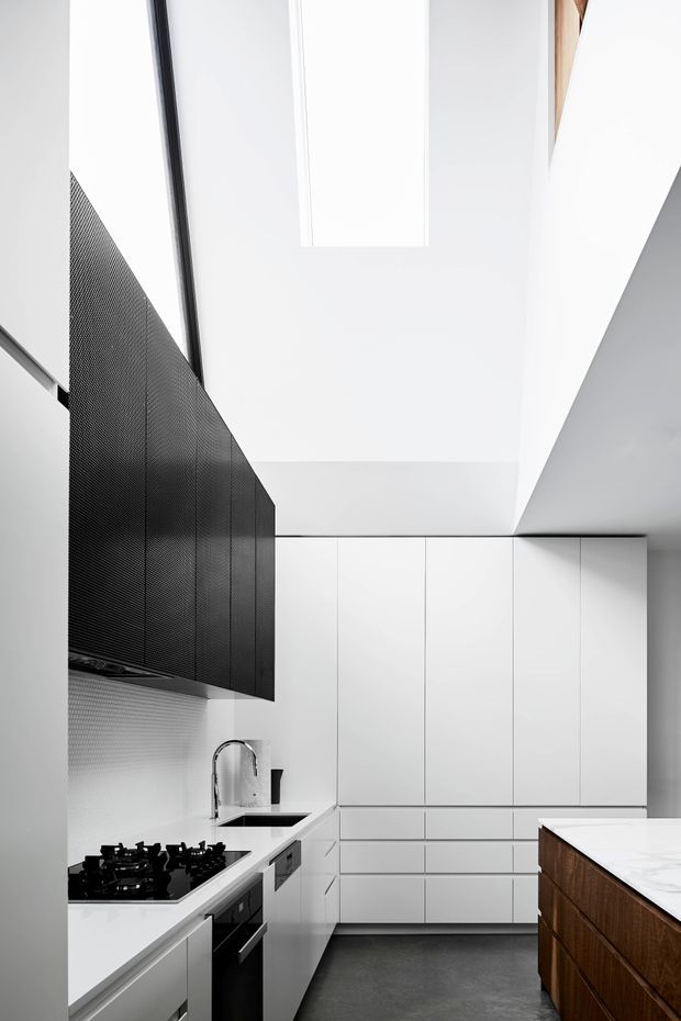 A narrow, highly streamlined modern kitchen viewed toward a wall of tall white flat-panel cabinetry. The space features a white benchtop with black gas cooktop and integrated oven, a stainless-steel gooseneck sink tap, dark textured overhead cabinetry, dark grey flooring, and a contrasting timber island or peninsula in the foreground. A large skylight or rooflight brings natural light into the tall white ceiling.