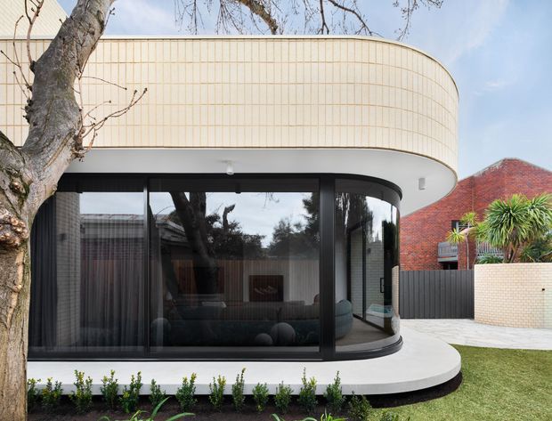 Exterior view of a contemporary curved building extension with a rounded upper-level facade clad in narrow cream-coloured tiles or panels. Full-height dark-framed glazing wraps around the lower level, revealing a living room inside. The structure sits beside a paved courtyard and landscaped garden, with a neighbouring red-brick building visible in the background.