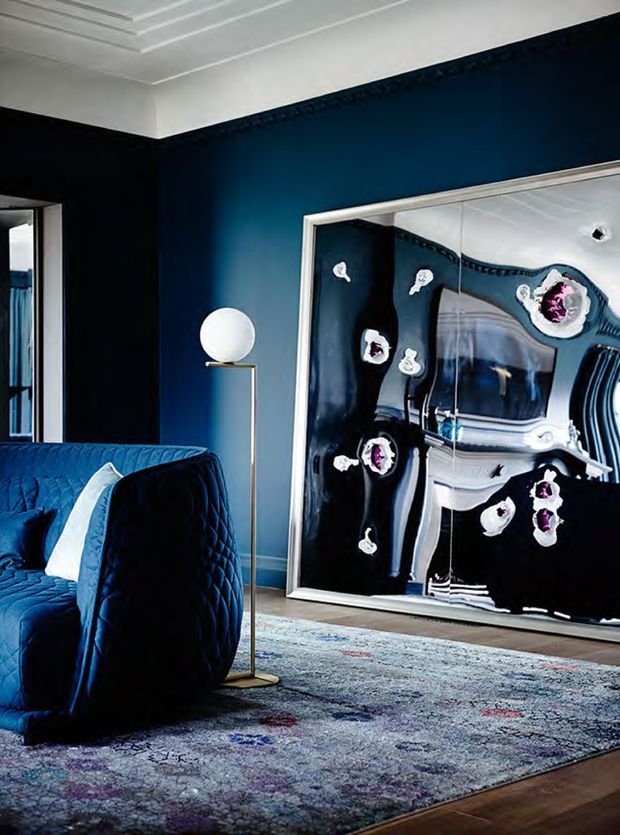 A dramatic, richly styled living room with deep navy walls, a dark blue quilted sofa, brass floor lamp, patterned area rug, and a large framed glossy black-and-white artwork leaning against the wall. The room features a white coffered ceiling and decorative dark trim, creating a luxurious, high-contrast atmosphere.