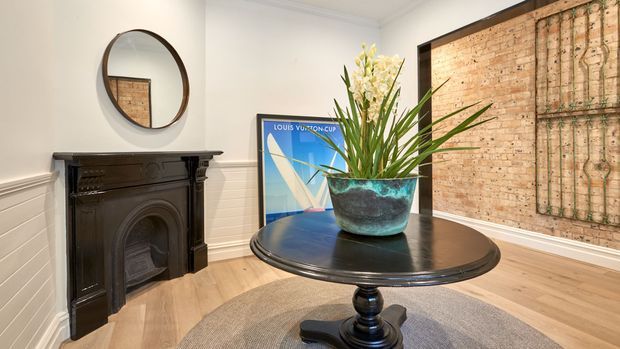 Elegant traditional interior corner featuring a dark carved fireplace mantel, round wall mirror, round pedestal table with a large potted plant, light timber flooring, white panelled walls, and a decorative framed artwork or textile panel.