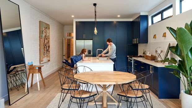 Bright open-plan kitchen and dining area with deep navy floor-to-ceiling cabinetry, a pale stone island, light timber flooring, and a round timber dining table surrounded by black wire-frame chairs. White-painted brick walls, high windows, pendant lighting, and a large indoor plant create a warm contemporary family space.