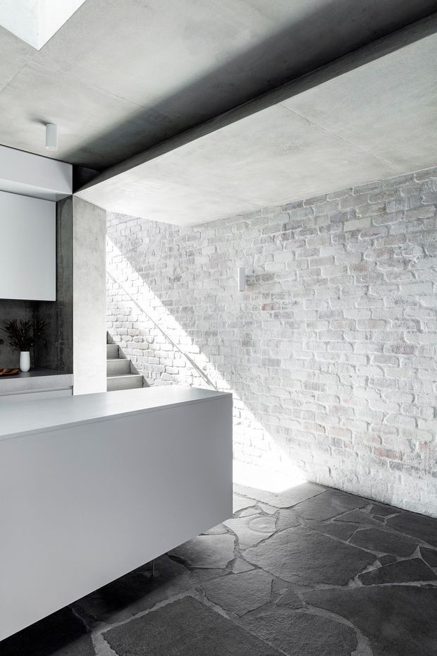 A stark, light-filled minimalist interior with a white-painted exposed brick wall, raw concrete ceiling and structural beams, dark irregular stone flooring, and a sleek white built-in island or counter. A small stairway is visible at the rear, with strong sunlight creating diagonal shadows across the brickwork. The space has an industrial, monolithic character with a restrained monochrome palette.