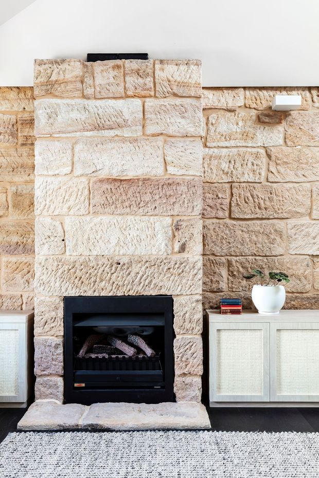 Interior detail of a traditional stone fireplace with a black firebox and broad rough-hewn masonry surround. Pale cream and warm beige stonework dominates the wall, with low light cabinetry on either side, a dark floor, and a textured neutral rug. Minimal decor includes books and a small potted plant.
