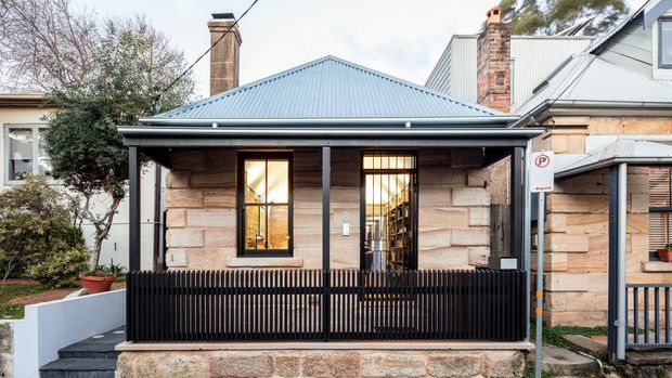 Street-facing view of a compact heritage-style cottage or terrace facade with a weathered timber-clad front porch, black vertical batten balustrade and posts, stone masonry base, tall timber-framed windows and door, and a blue-grey corrugated metal gabled roof. Adjacent attached or neighbouring structures are visible at either side.