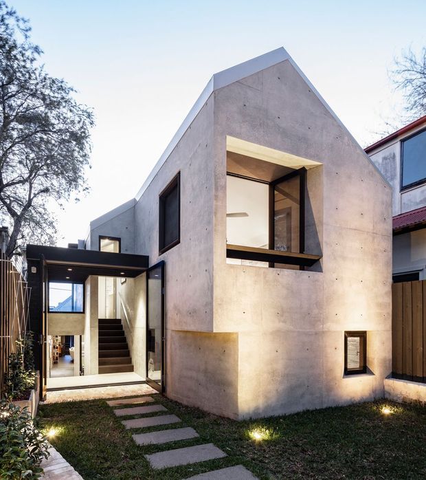 Exterior view of a compact contemporary residence with a sharply pitched gable roof form, monolithic exposed concrete walls, dark-framed glazing, and a recessed entry. A landscaped garden path leads toward the illuminated interior and stairway, with a neighboring dwelling visible at the right.