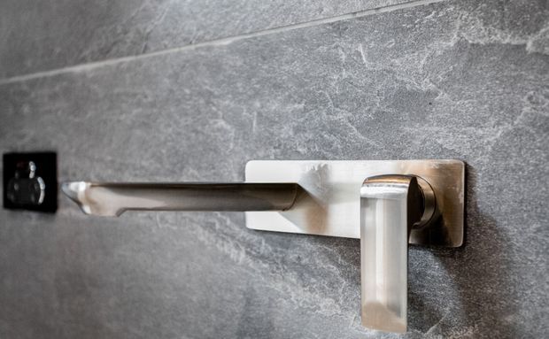 Close-up of a contemporary wall-mounted metal tap or control fixture against a dark grey textured tiled wall, with a small black wall switch visible to the left.
