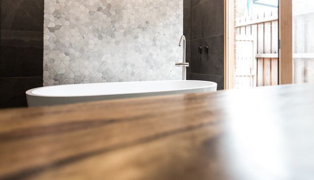 A contemporary bathroom featuring a freestanding white bathtub against a wall of pale irregular hexagonal tiles, framed by dark charcoal wall tiles. A slim wall-mounted bath filler and hand shower are visible beside the tub, with a timber-framed opening and vertical timber screening at right. A broad warm timber surface occupies the foreground.