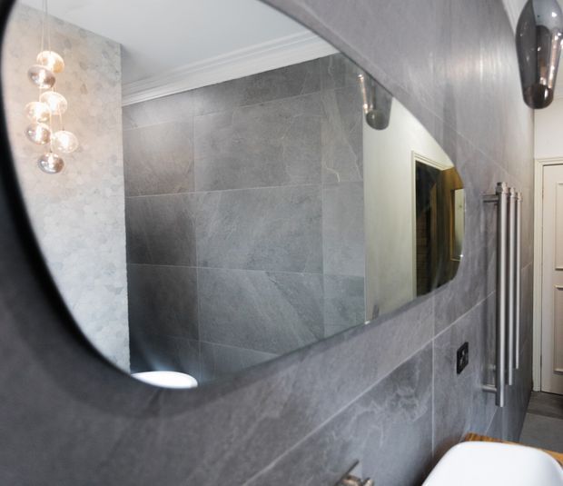 Close-up view of a refined bathroom featuring large-format grey stone-look wall tiles, a broad rounded rectangular mirror, sculptural pendant lighting reflected in the mirror, a white basin edge, and tall brushed-metal towel rails. The space has a cool, polished, contemporary feel with decorative ceiling moulding and pale patterned wallcovering visible at the side.