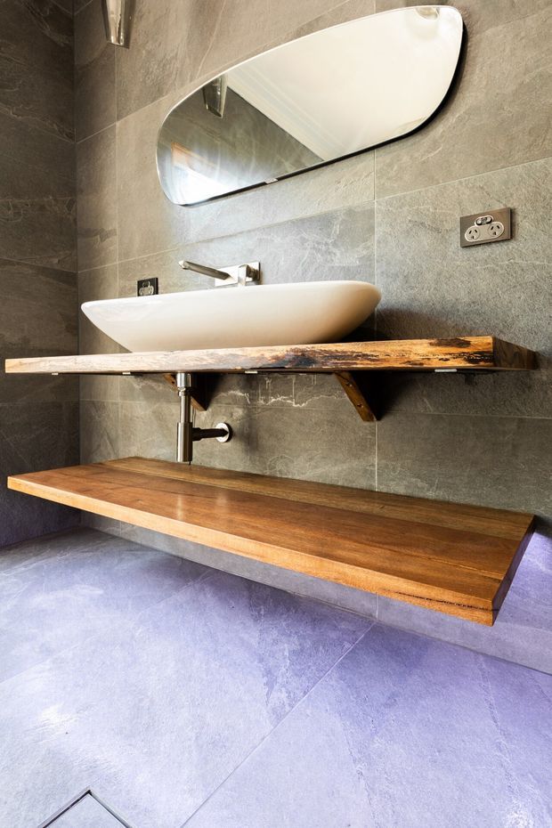 A contemporary bathroom vanity featuring a white vessel basin, wall-mounted chrome tapware, an organically shaped mirror, and two substantial reclaimed timber shelves. The walls and floor are finished in large-format textured grey stone-look tiles, with exposed plumbing and a double power outlet adding an industrial character.