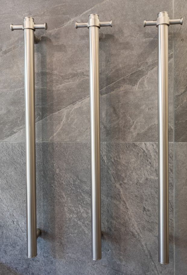 Close-up of three tall brushed metal vertical rails or handles mounted against a dark grey natural-stone tiled wall. The image focuses on bathroom-style hardware and textured stone finishes, with a clean, neutral, contemporary appearance.