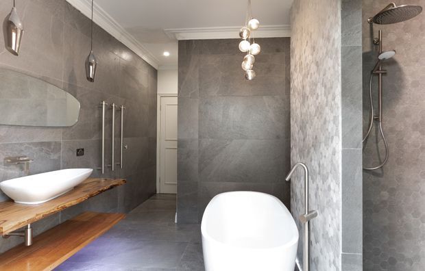 Contemporary bathroom featuring a freestanding white bathtub, wall-mounted rainfall shower, floating timber vanity with vessel basin, large grey stone-look wall and floor tiles, a mosaic-tiled shower partition, sculptural pendant lighting, and polished metal fixtures.
