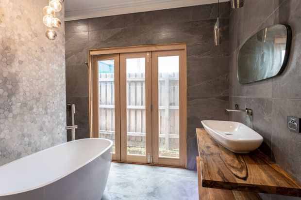 A contemporary bathroom featuring a freestanding white bathtub, a floating timber vanity with a vessel basin, dark grey stone-look wall tiles, a pale patterned feature wall, timber-framed glazed French doors, and a rounded rectangular mirror. Globe pendant lighting adds a warm decorative accent.