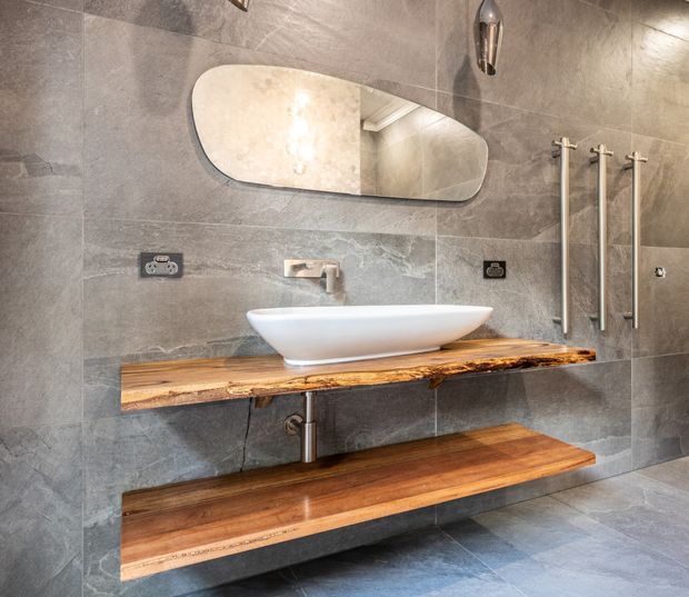 Contemporary bathroom vanity with a white vessel basin on a thick live-edge timber benchtop, paired with a matching floating timber shelf. Large-format grey stone-look wall and floor tiles, an organically shaped mirror, brushed metal tapware, and vertical heated towel rails create a refined, warm modern interior.