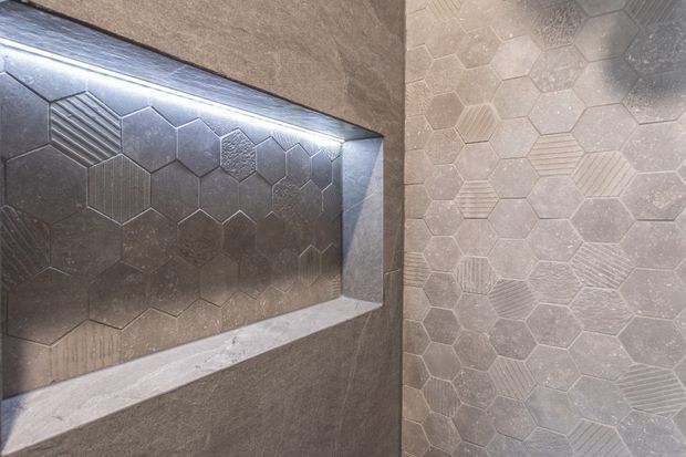 Close-up of a tiled wall and recessed niche featuring a mix of hexagonal and elongated hexagonal tiles with varied embossed linear and textured patterns. The surfaces are finished in coordinated dark and medium grey tones, with concealed lighting illuminating the niche.
