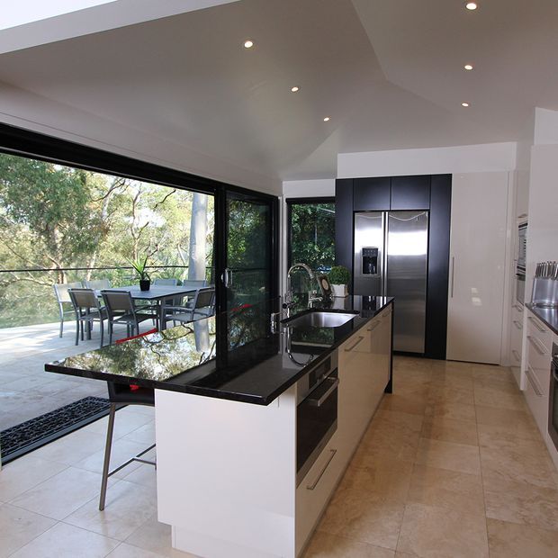 A bright, contemporary kitchen with a central island featuring a glossy black stone benchtop, integrated sink and bar seating. White cabinetry, dark upper cupboards, and stainless-steel appliances line the rear wall. Full-height black-framed glazing opens the kitchen to a paved outdoor dining terrace and leafy surroundings. Recessed ceiling lighting and pale stone-look flooring complete the clean, modern space.
