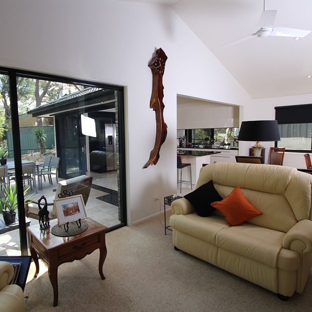 Bright open-plan living room with a cream upholstered sofa, dark and orange cushions, traditional wooden side furniture, table lamp, wall-mounted carved timber artwork, and beige carpet. Full-height glazing opens to a tiled patio and enclosed outdoor area, while the adjoining kitchen and dining space are visible in the background. A white ceiling fan and recessed lighting complete the room.