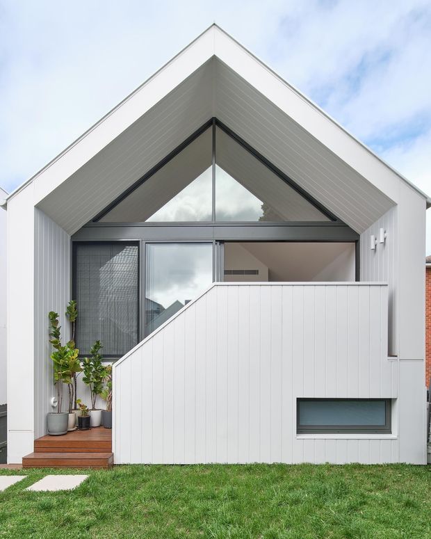 Front exterior view of a compact contemporary house with a prominent gabled roof and white vertical cladding. The facade features a large triangular glazed section beneath the gable, dark window and door framing, a sheltered entry with timber decking and potted plants, and a white privacy screen enclosing the front area.