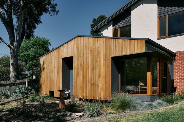 Exterior view of a contemporary residential extension with a low-pitched dark roof and warm vertical timber cladding. The addition features deep, dark-framed openings and large timber-framed glazing, attached to a taller existing brick-and-render house. Native-style planting, grasses, mature trees, and a timber boundary fence surround the building.