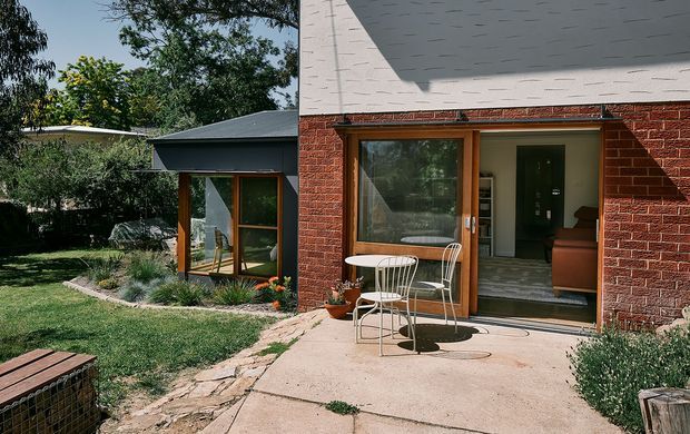 Exterior view of a low-rise brick house with a dark low-pitched roof, large timber-framed glazed doors and windows, and a small concrete patio furnished with a white metal café table and chairs. The landscaped garden includes lawn, planting beds, mature trees, and a stone-paved path. Interior living space is visible through the glazing.