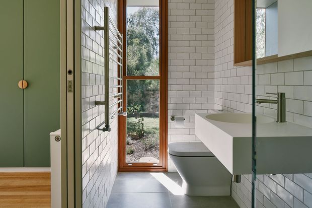 A contemporary bathroom viewed from the doorway, featuring white subway-tile walls, a wall-mounted white vanity with integrated basin, chrome tapware, a wall-hung toilet, a heated towel rail, dark grey floor tiles, and a full-height timber-framed window overlooking greenery. A muted green door and warm timber detailing add contrast.